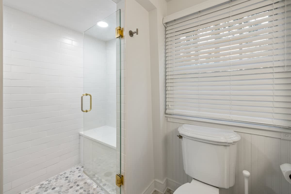 Modern bathroom with a walk-in shower featuring white subway tiles, a glass door with gold hardware, and a built-in bench. The floor has small hexagonal tiles, and a toilet is visible next to a window with blinds.