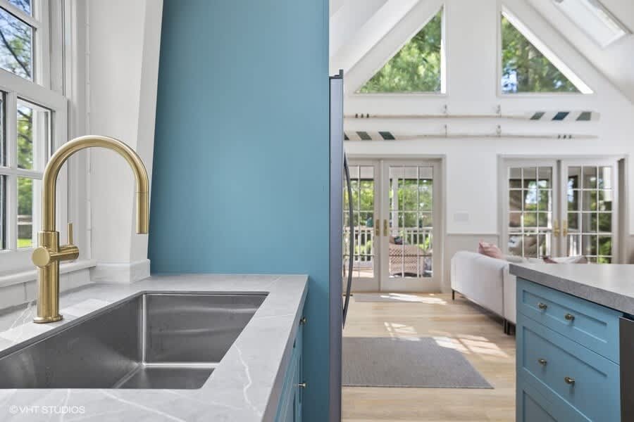 Modern kitchen corner with a deep stainless steel sink and a gold gooseneck faucet, set against a blue accent wall and light gray countertops, with a view into the living area through French doors.