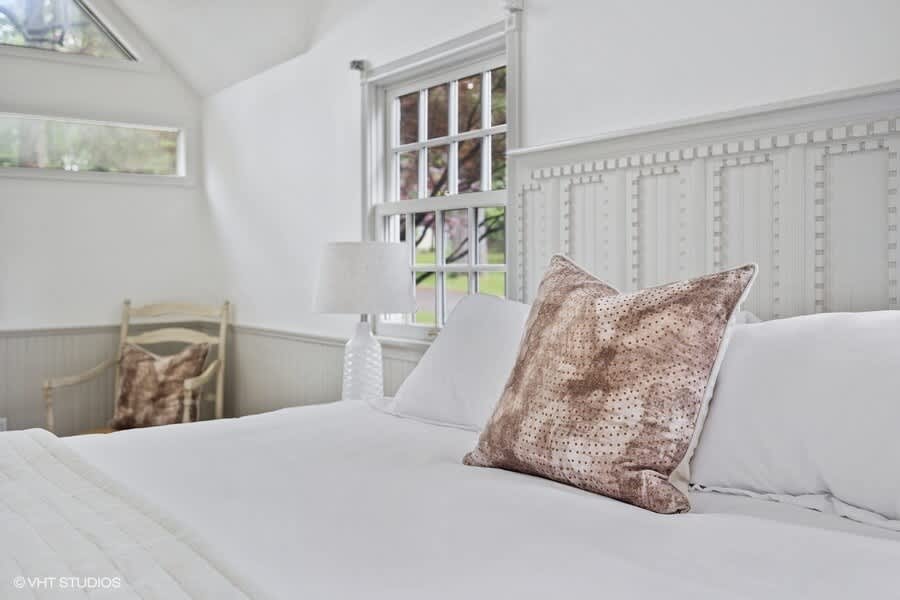 Serene bedroom (Bedroom-1) featuring a white bed with a tall, light gray paneled headboard, a patterned throw pillow, and a white nightstand with a lamp beside a multi-pane window.
