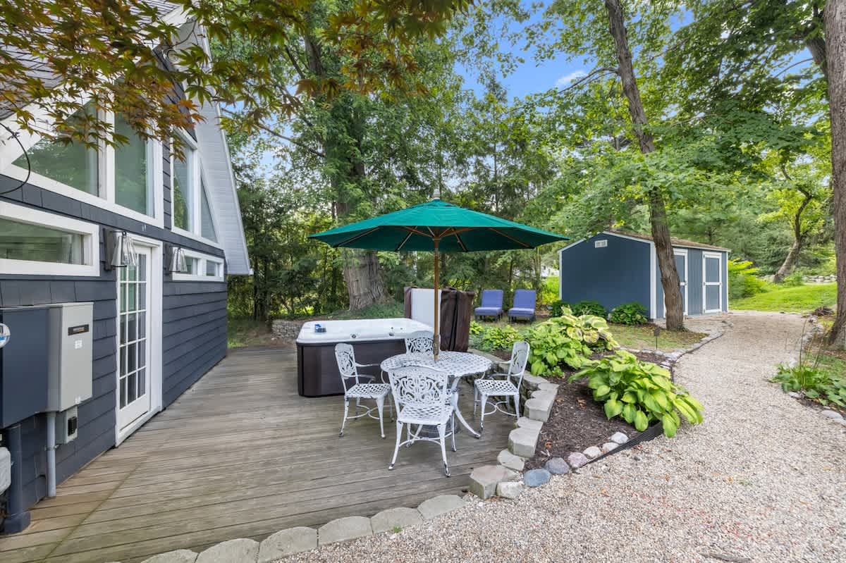 A spacious wooden deck features a white hot tub and a matching white metal outdoor dining set with an umbrella, positioned next to the dark-sided house with large windows and surrounded by lush green trees.