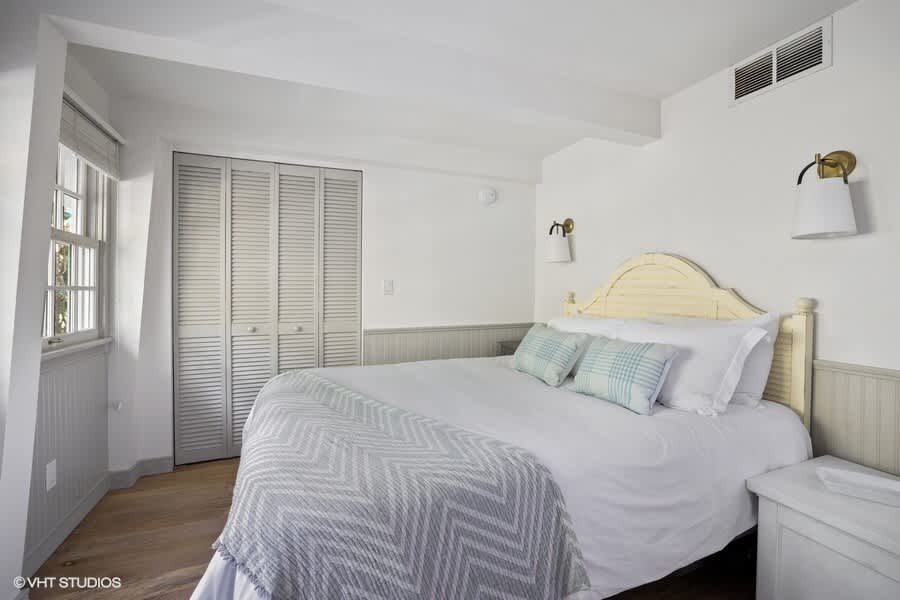 Cozy bedroom featuring a queen bed with white bedding and blue accent pillows, light wood floors, white wainscoting, and louvered closet doors. A window with blinds provides natural light.