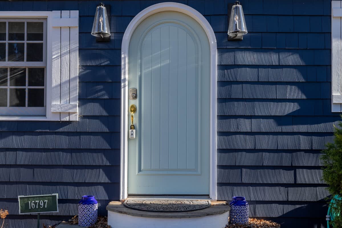 Close-up of a light blue arched front door with brass hardware, flanked by white shutters and two outdoor sconces, set against dark blue shingle siding.