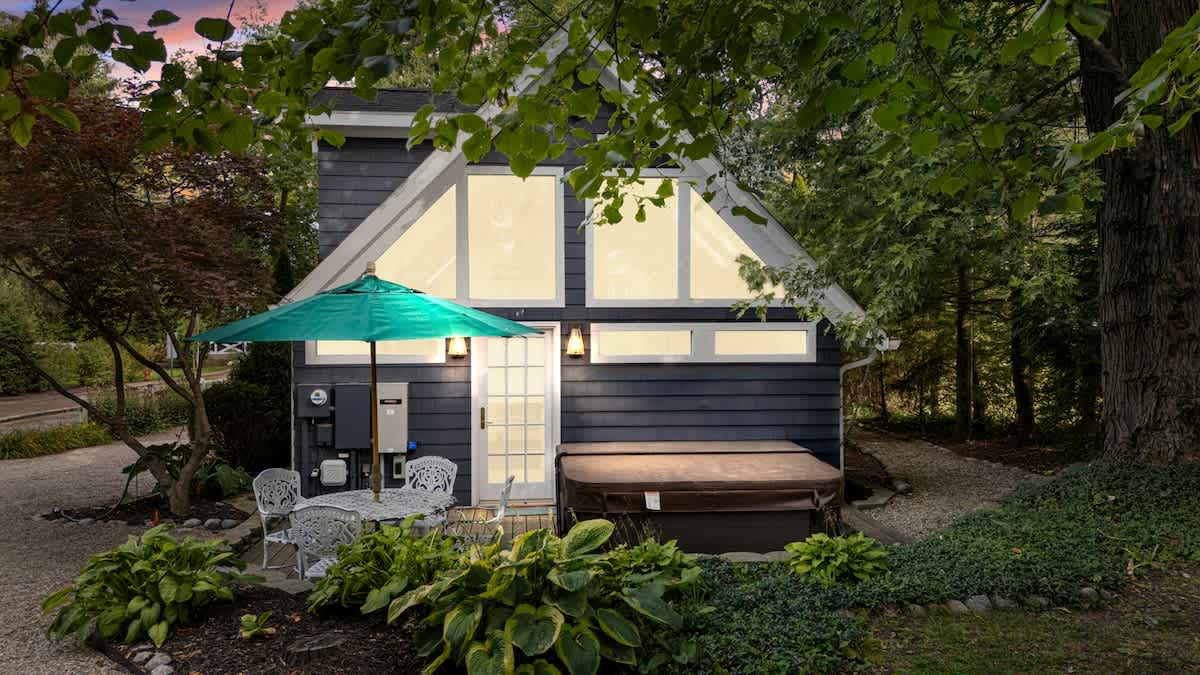 Exterior view of a modern structure with large windows, featuring a hot tub on a gravel patio. An outdoor dining set with an umbrella is also present, surrounded by lush greenery.