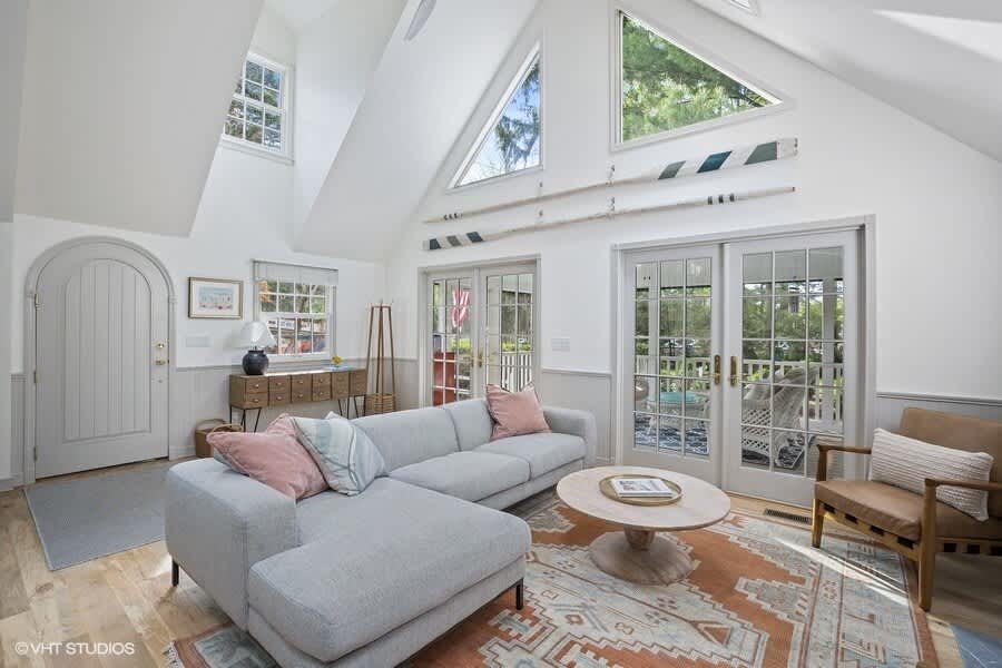 Inviting living room with a light gray sectional sofa and a round wooden coffee table on an ornate rug, featuring a unique arched front door, large triangular windows, and French doors leading to an outdoor space.