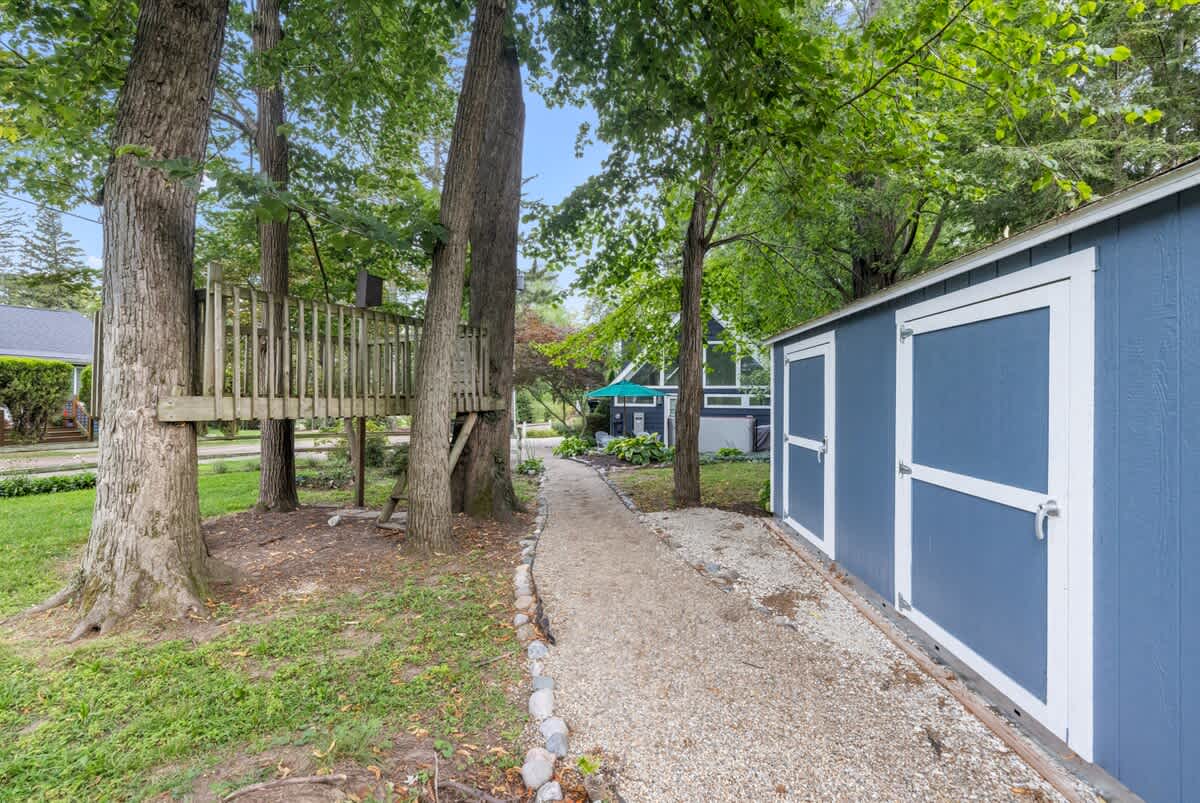 A gravel path winds through a verdant yard, leading past a blue shed with white trim and a rustic wooden treehouse nestled among tall, mature trees.