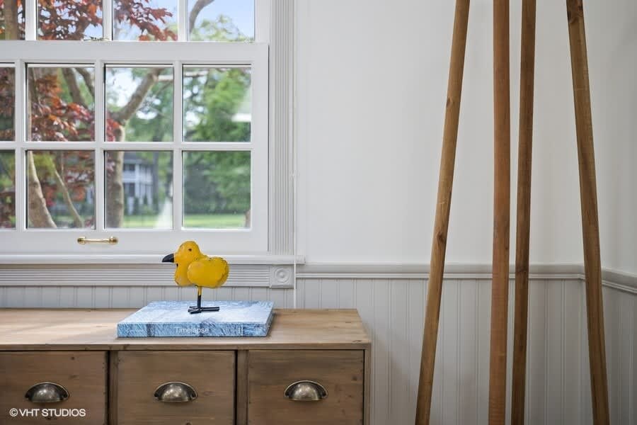 Close-up of a wooden console table with drawers and metal pulls, featuring a yellow decorative duck on a blue book, positioned next to a large window with white grilles and a wooden coat rack.