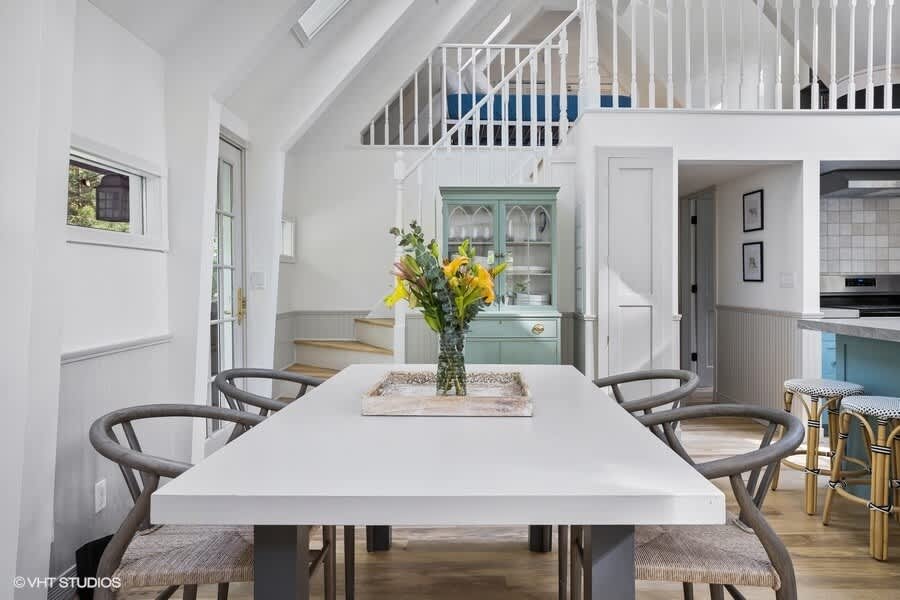 Elegant dining area with a white rectangular table and six woven-back chairs, set against a backdrop of a light blue hutch and a staircase leading to a loft, with a vase of flowers as a centerpiece.