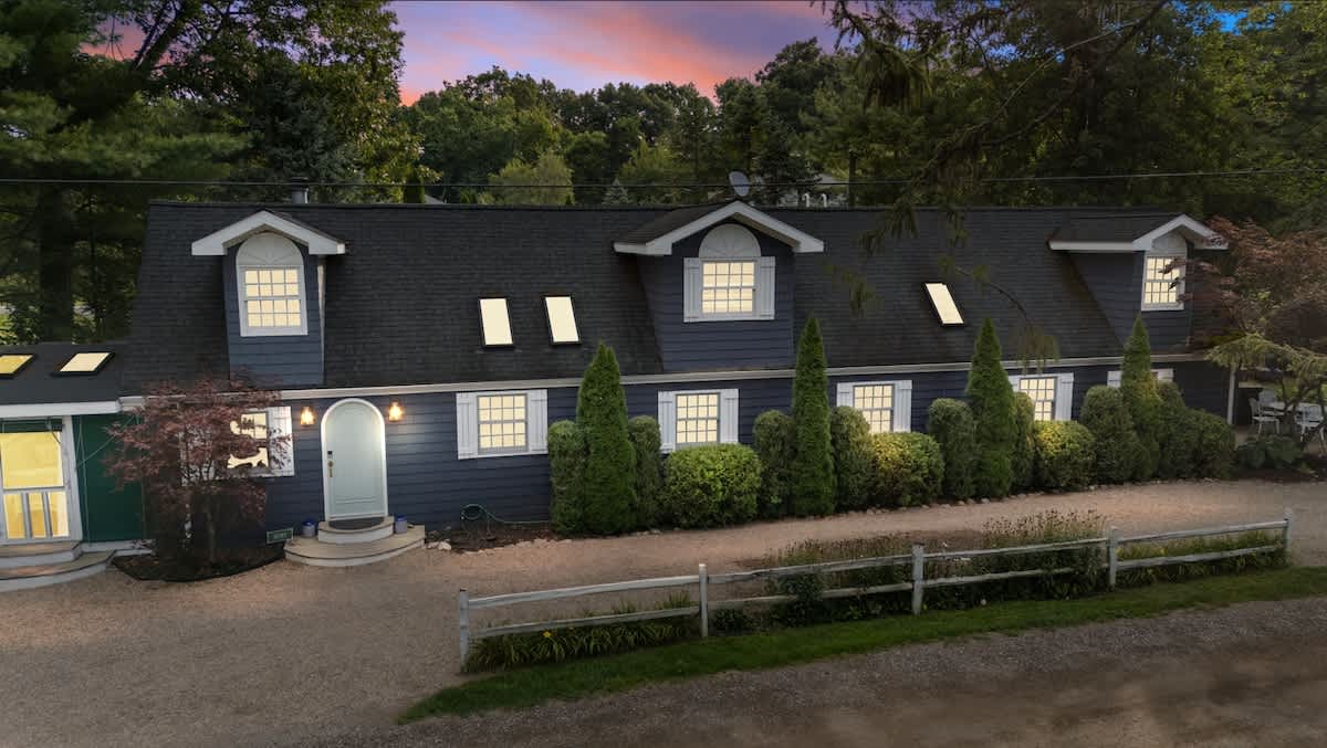 Charming dark blue house with white trim and multiple dormer windows, illuminated at dusk. The property features a gravel driveway, manicured shrubs, and a welcoming front entrance.