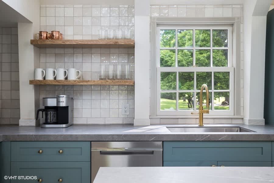 Stylish kitchen area with open wooden shelving displaying mugs and glasses, a white tiled backsplash, and a large window above a stainless steel sink with a gold faucet, flanked by blue cabinets.