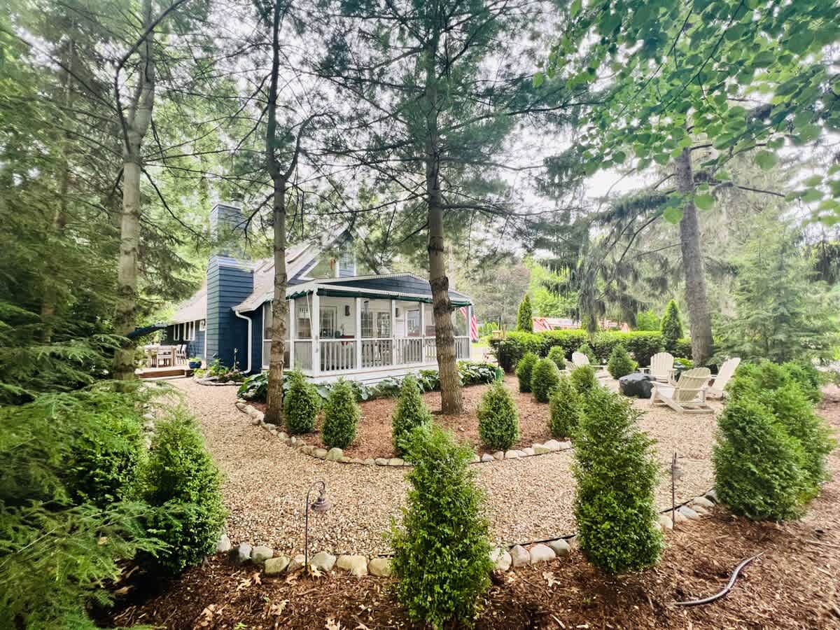Lush backyard featuring a gravel path leading to a fire pit area with Adirondack chairs, surrounded by manicured shrubs and tall trees. A covered porch of the house is visible in the background.