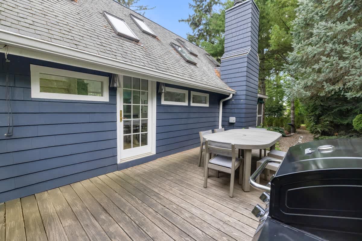Expansive wooden deck with an outdoor dining set and a BBQ grill, adjacent to the dark blue house with a glass-paned door and multiple windows. Tall trees and greenery provide privacy.