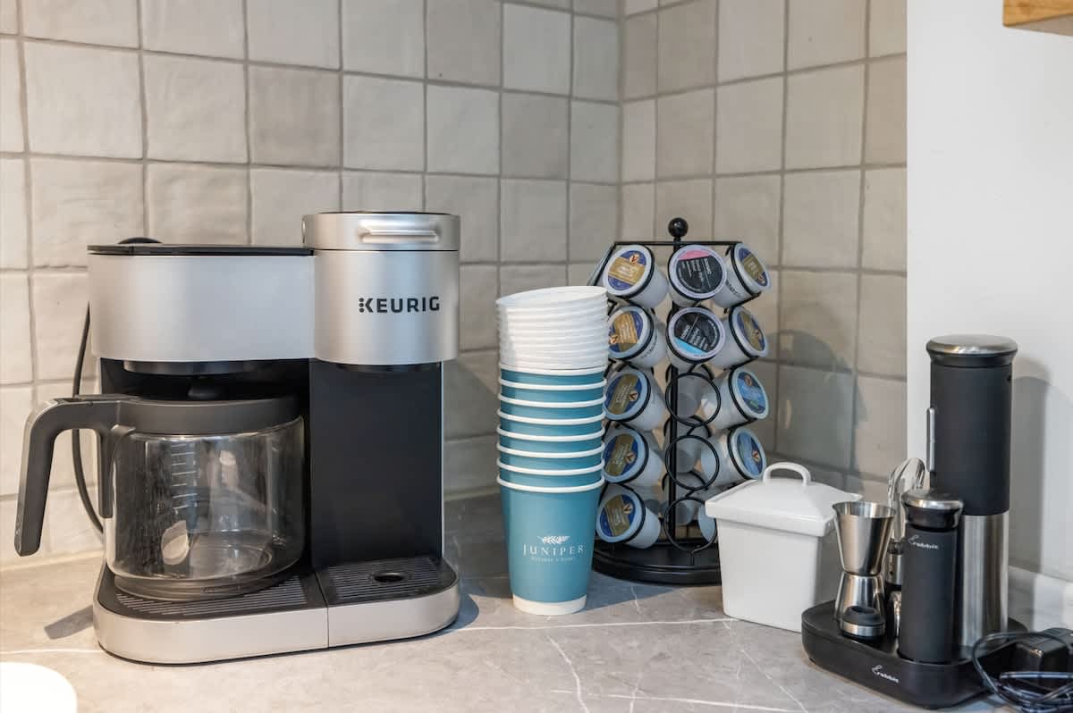 Dedicated coffee station on a light gray countertop with a white tiled backsplash, featuring a Keurig coffee maker, a stack of paper cups, a K-cup carousel, and various coffee accessories.