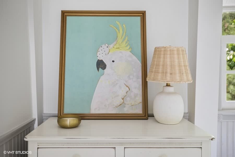 A framed painting of a white cockatoo with a yellow crest, displayed on a white dresser next to a white ceramic lamp with a woven shade, adding a decorative touch.