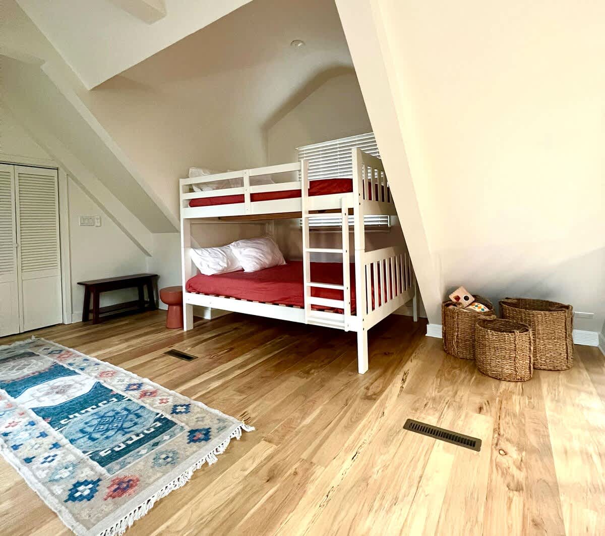 Bright bedroom with white wooden bunk beds featuring red bedding, light hardwood floors, and a colorful patterned rug. The room has sloped ceilings and wicker baskets for storage.