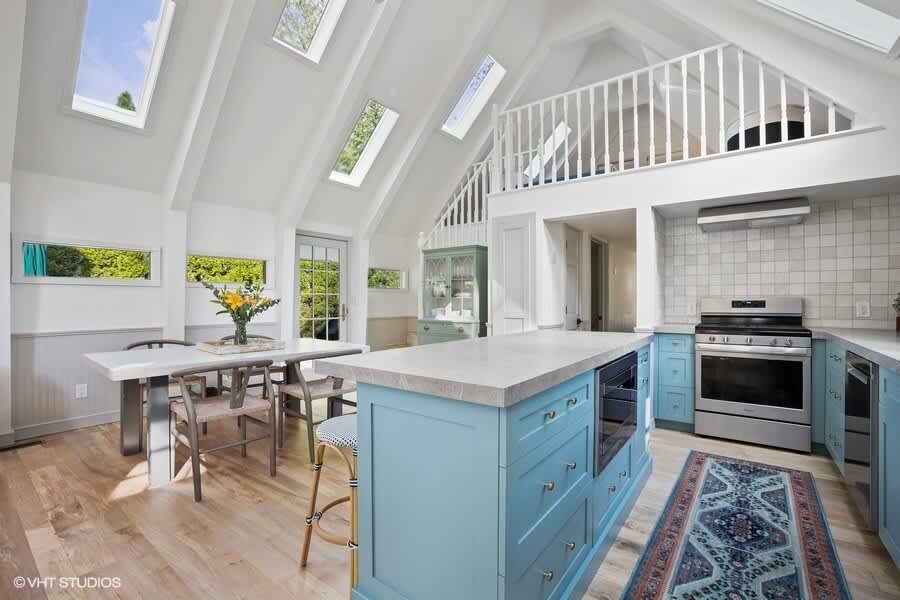 Bright open-concept kitchen and dining area with a vaulted ceiling and skylights, featuring a large blue kitchen island with bar stools, a white dining table with chairs, and light wood floors.