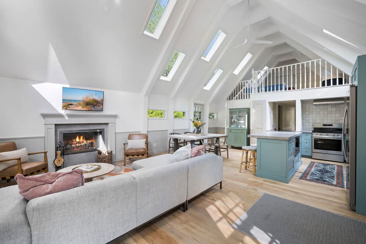 Bright, open-concept living space with a high vaulted ceiling, multiple skylights, and light wood floors, featuring a gray sectional sofa, a dining area, and a blue kitchen island, with a loft visible above.
