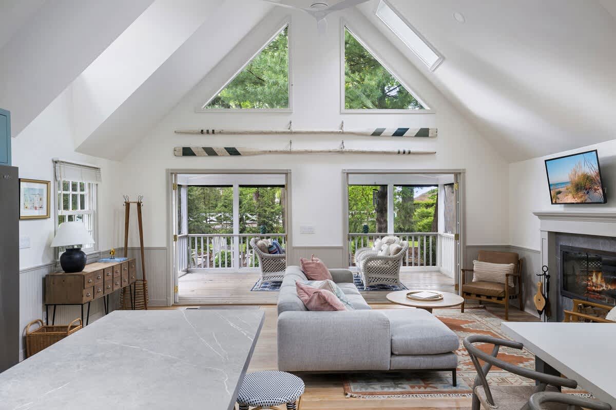 Spacious living area with a vaulted ceiling and large triangular windows overlooking trees, featuring a gray sectional sofa, a round coffee table, and two decorative oars mounted on the wall, with French doors leading to a deck.