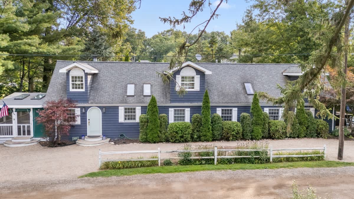 An aerial view captures the long, dark blue house with a gray shingled roof, multiple dormer windows, and white trim, set amidst a landscape of mature trees and a wide gravel driveway.