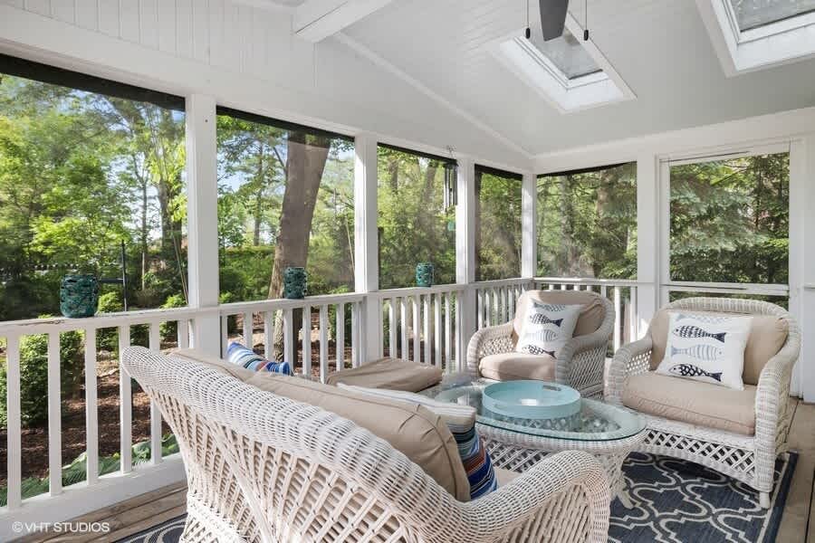 Screened porch with white wicker sofa and armchairs, a glass-top coffee table, and a blue patterned rug, offering views of a lush wooded area. The ceiling features skylights and a fan.