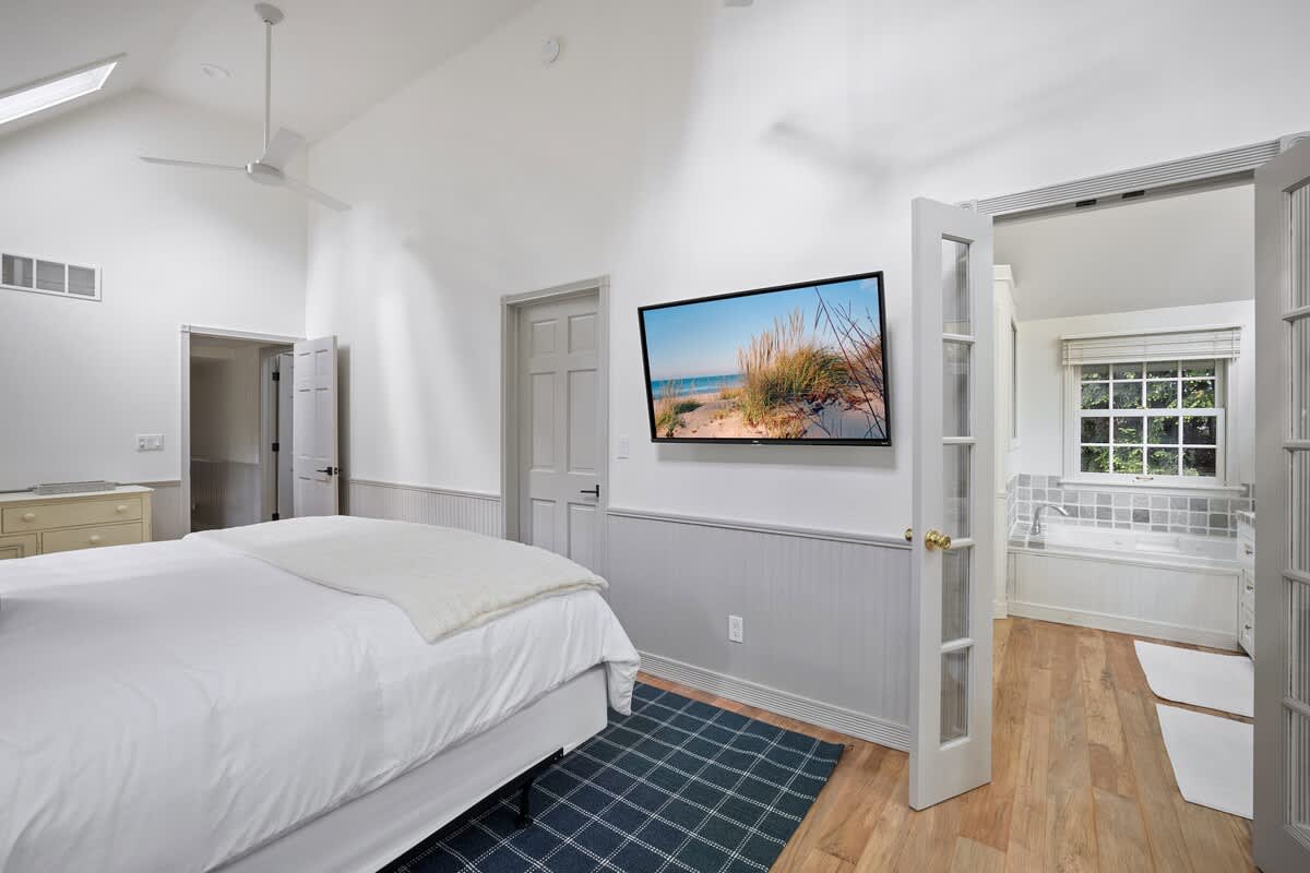 Master bedroom (Bedroom-1) with a white bed, a large wall-mounted TV displaying a beach scene, and light gray wainscoting. French doors on the right open to an en-suite bathroom with a visible tub.