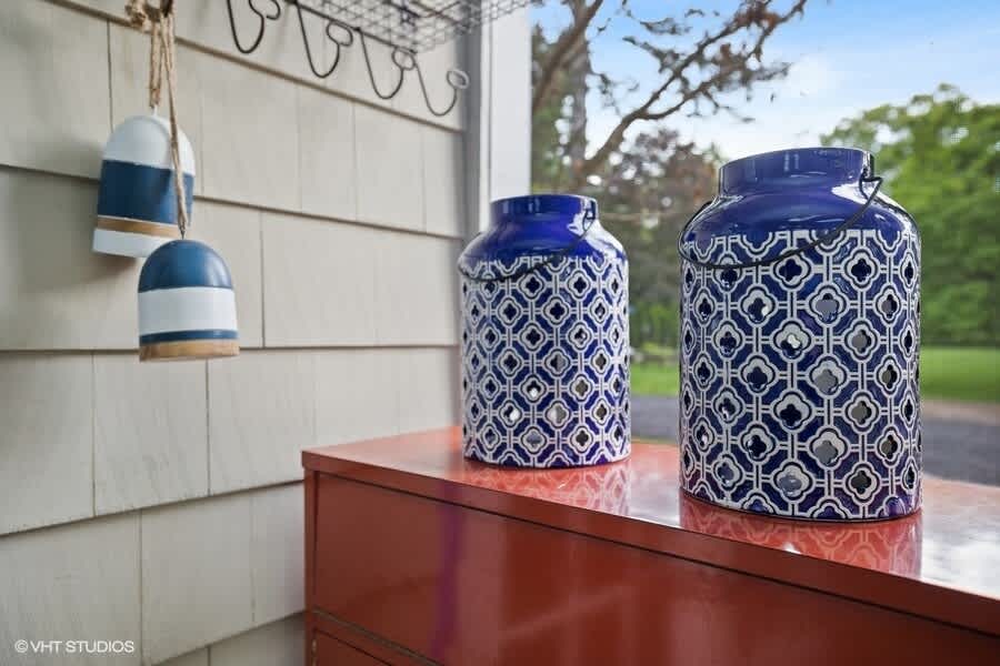Close-up of two blue and white patterned ceramic lanterns on a reddish-brown console table, set against a light-colored shingled exterior wall with decorative buoys hanging nearby.
