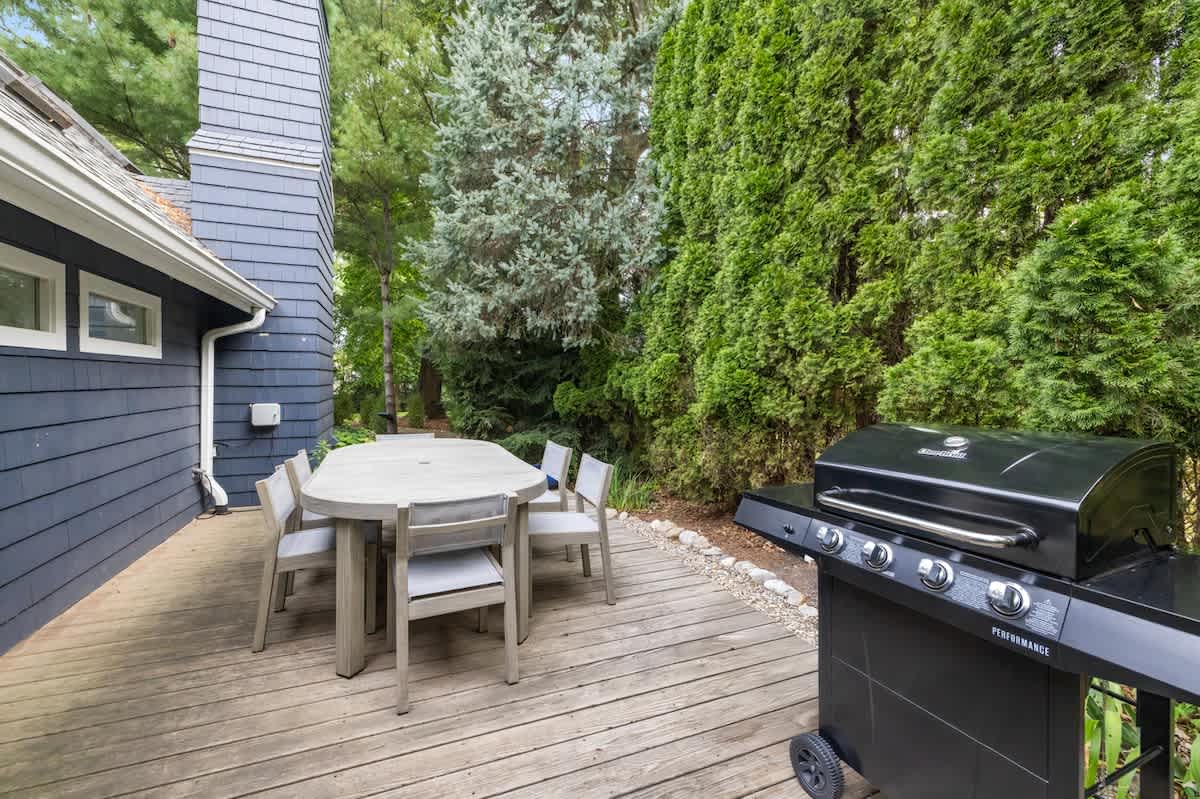 Spacious wooden deck featuring an outdoor dining table with chairs and a black BBQ grill, set against the dark blue siding of the house. Surrounded by tall green hedges.