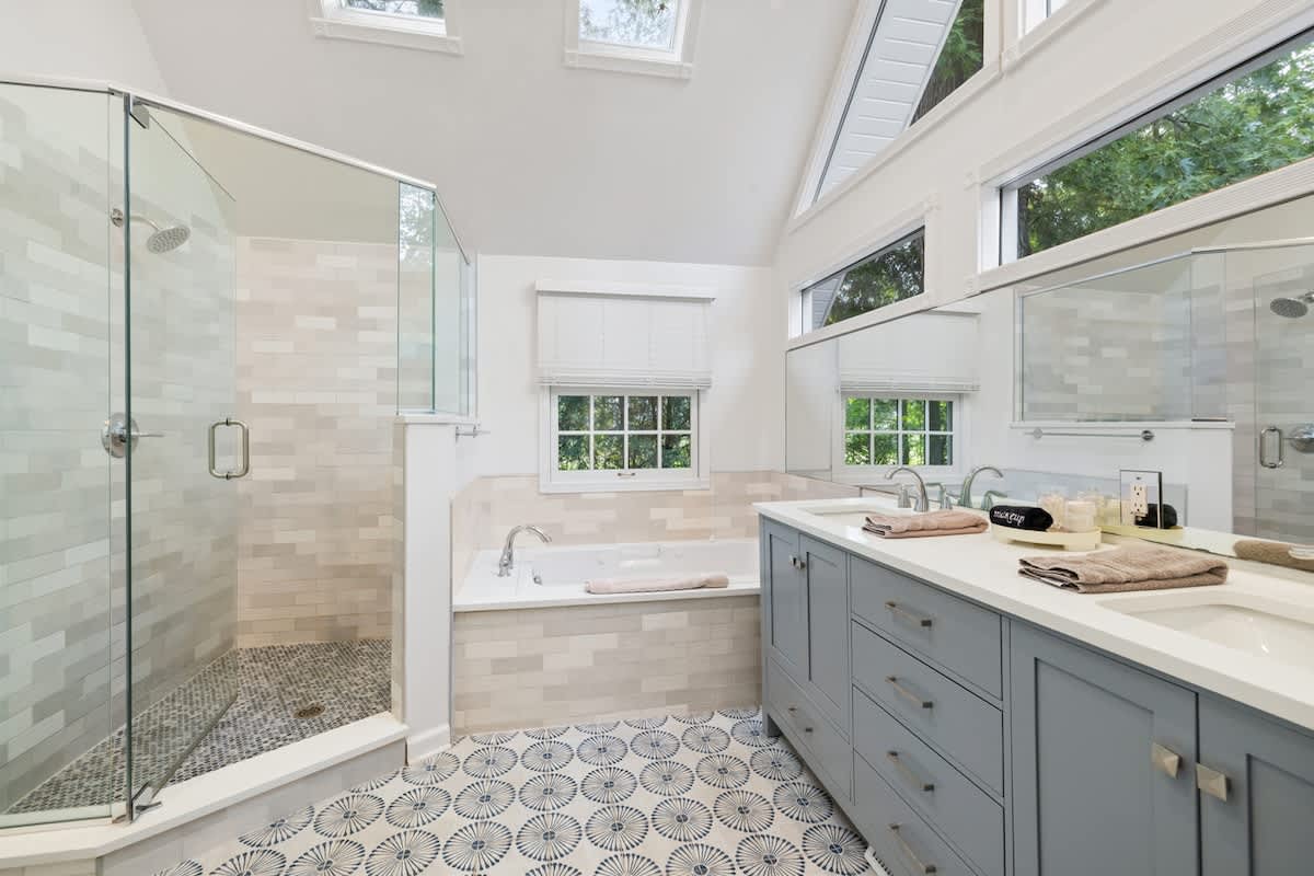 Spacious and bright bathroom (Bathroom-1) featuring a glass-enclosed shower, a built-in bathtub, and a double vanity with light blue cabinets and white countertops. Blue and white patterned floor tiles add a decorative touch, complemented by skylights.