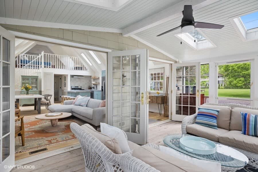 Bright living room with a white wicker sofa and armchair, a glass coffee table, and a ceiling fan, connected to an open-concept dining area and kitchen through French doors. Skylights and light hardwood floors enhance the airy feel.