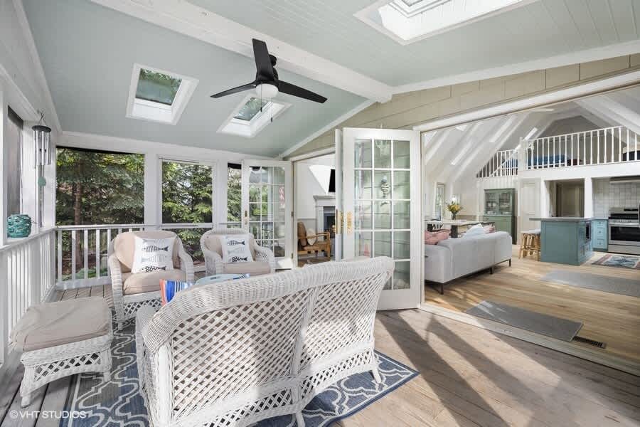 Screened-in porch with light blue ceiling, wicker seating, skylights, and French doors opening into the main living area, offering a bright and airy outdoor-indoor transition.