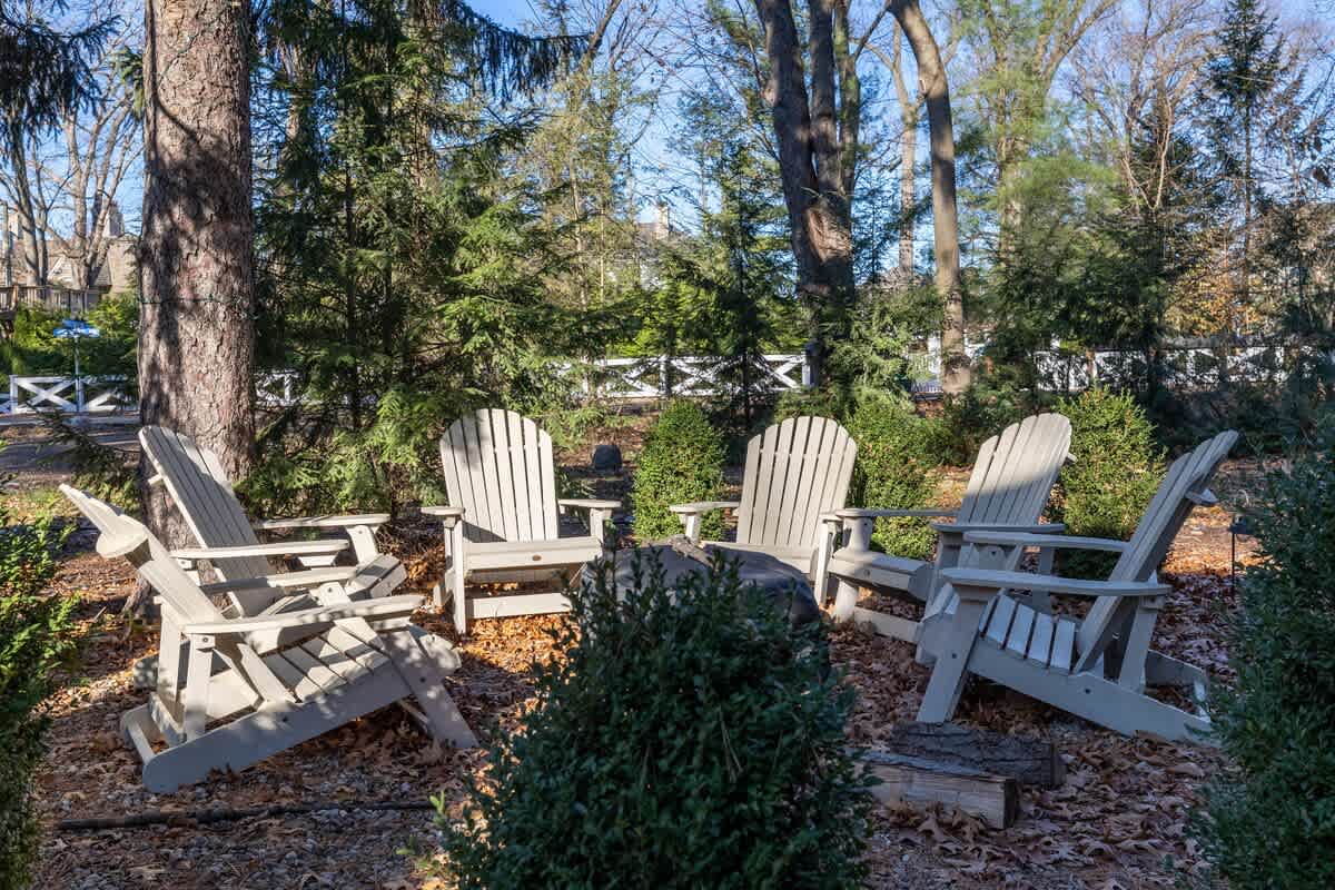 A cozy outdoor fire pit area features five light-colored Adirondack chairs arranged in a circle, surrounded by lush evergreen bushes and tall trees, creating a secluded gathering spot.