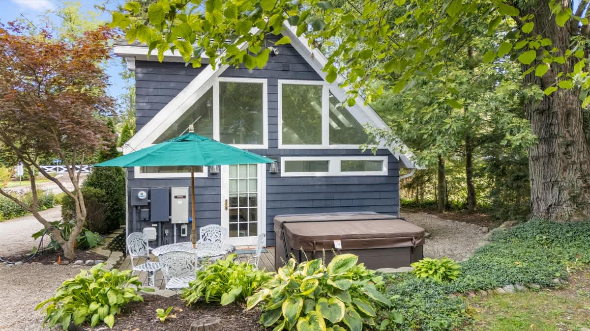 Exterior view of a dark-sided house with an A-frame window design, featuring an outdoor hot tub, a white patio table with chairs, and a green umbrella, surrounded by lush landscaping.