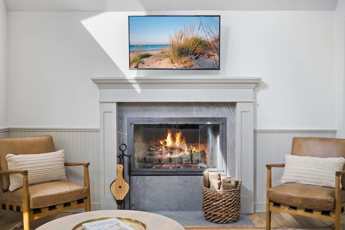Cozy living room with a white fireplace mantel, gray marble surround, and a roaring fire, flanked by two leather armchairs with textured pillows, under a wall-mounted TV displaying a beach scene.