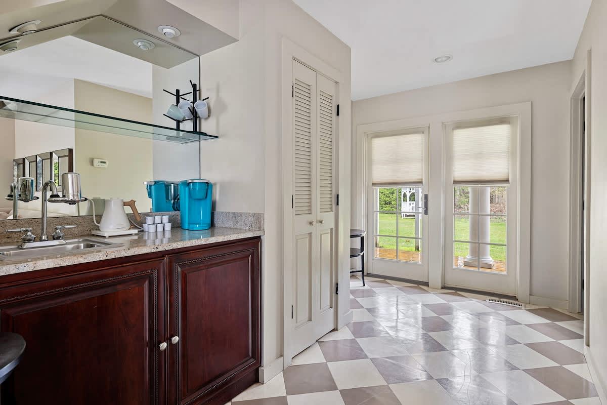 A wet bar area with dark wood cabinetry, granite countertops, and a mirrored backsplash, equipped with a sink and coffee makers. French doors with blinds open to an outdoor view, and the floor features a gray and white checkered pattern.