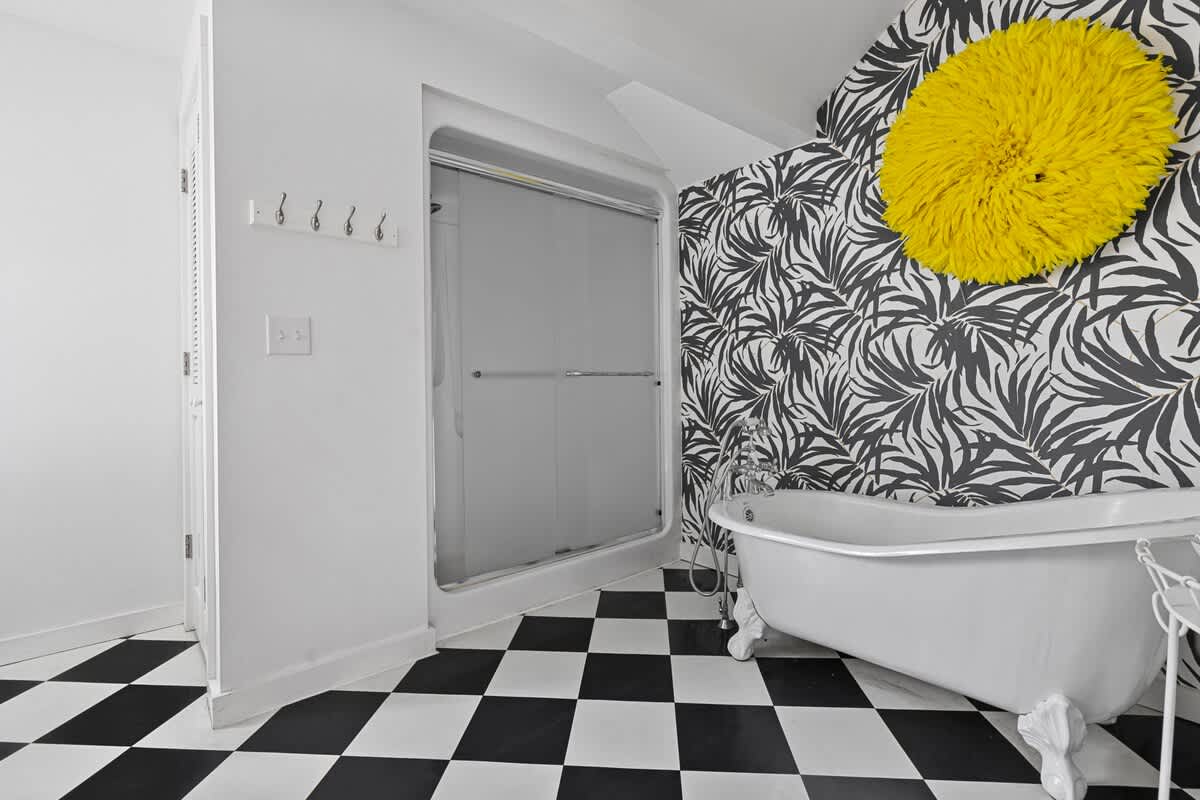 A vibrant bathroom with a bold black and white checkered floor, a classic white clawfoot tub set against a black and white tropical leaf wallpaper, and a bright yellow decorative wall hanging. A glass-enclosed shower is also visible.