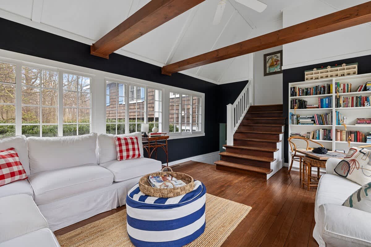 Inviting living room with white slipcovered sofas, dark blue walls, exposed wooden ceiling beams, and a wooden staircase, featuring large windows and a striped ottoman.