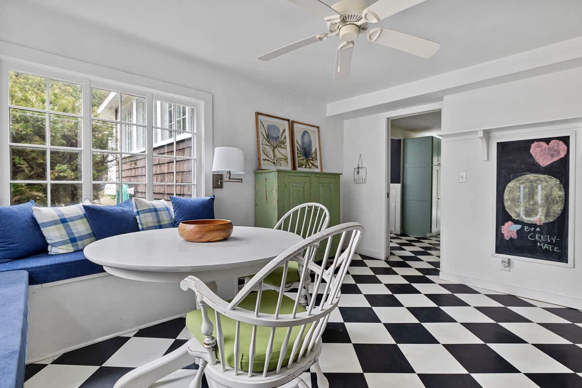 Another view of the breakfast nook, showing the round white table, built-in bench with blue cushions, and white chairs. A green cabinet and a chalkboard are visible on the wall, complementing the black and white checkered floor.