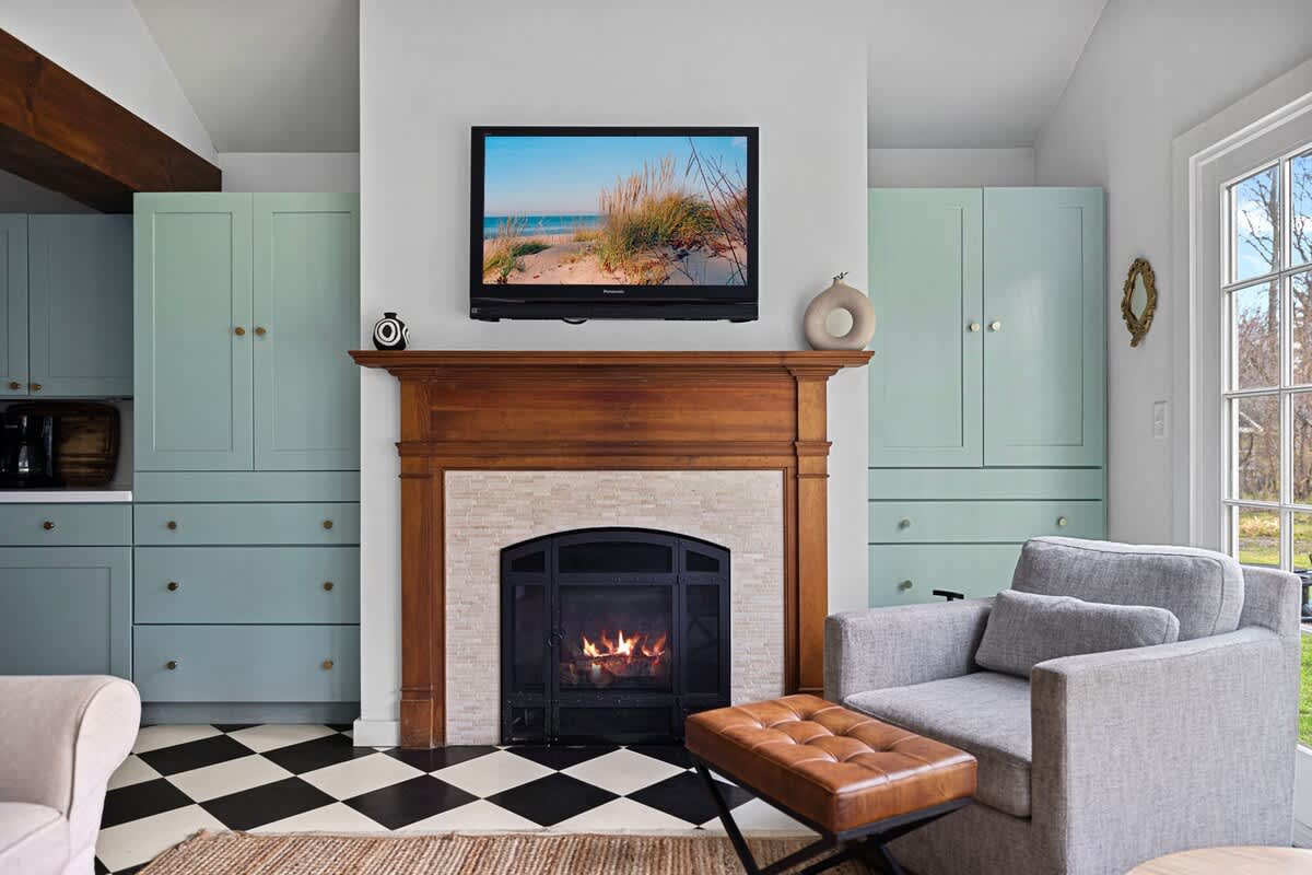 A cozy living area features a wooden mantel fireplace with a mounted TV above, flanked by light green built-in cabinets. A gray armchair with a leather ottoman sits on a black and white checkered floor, next to a large window.