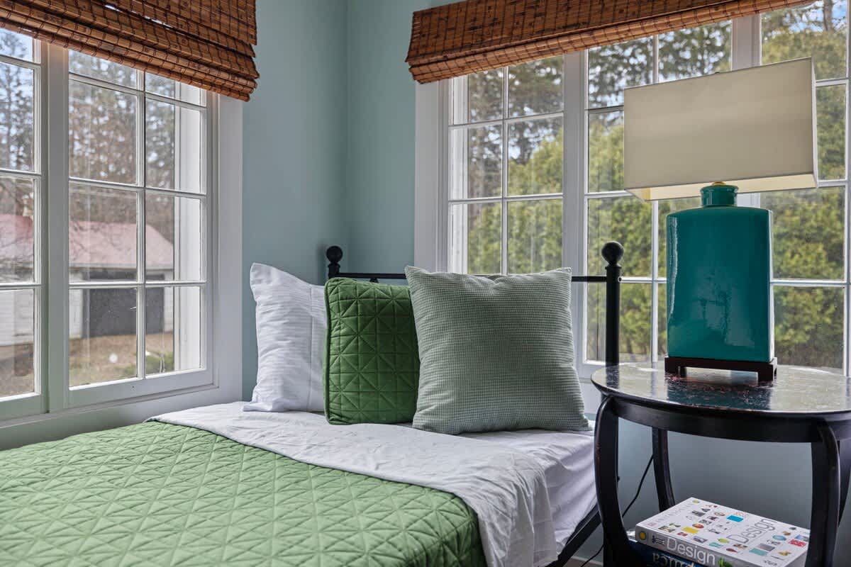 A close-up view of one twin bed in the bedroom, featuring green bedding and pillows, positioned next to a round black side table with a large turquoise lamp. Large windows with bamboo blinds provide natural light.