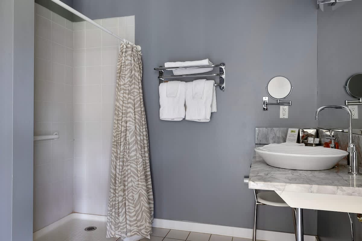 A modern bathroom featuring a walk-in shower with white subway tiles and a zebra-print shower curtain, alongside a marble-topped vanity with a white vessel sink and a wall-mounted mirror. The walls are painted a cool gray.