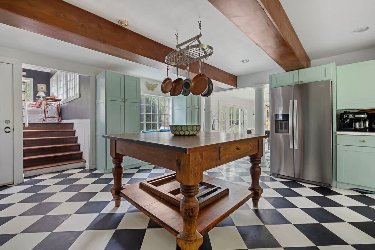 The kitchen features light green cabinetry, a large stainless steel refrigerator, and a central wooden island with a pot rack. The black and white checkered floor extends throughout, with a view towards a wooden staircase leading to another level.