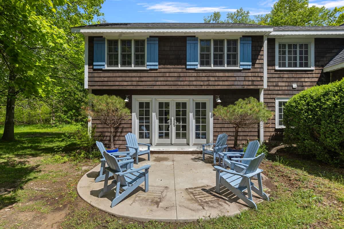 A concrete circular patio with light blue Adirondack chairs is situated at the back of a shingle-sided house, featuring French doors and blue window shutters. Lush greenery and trees surround the area.