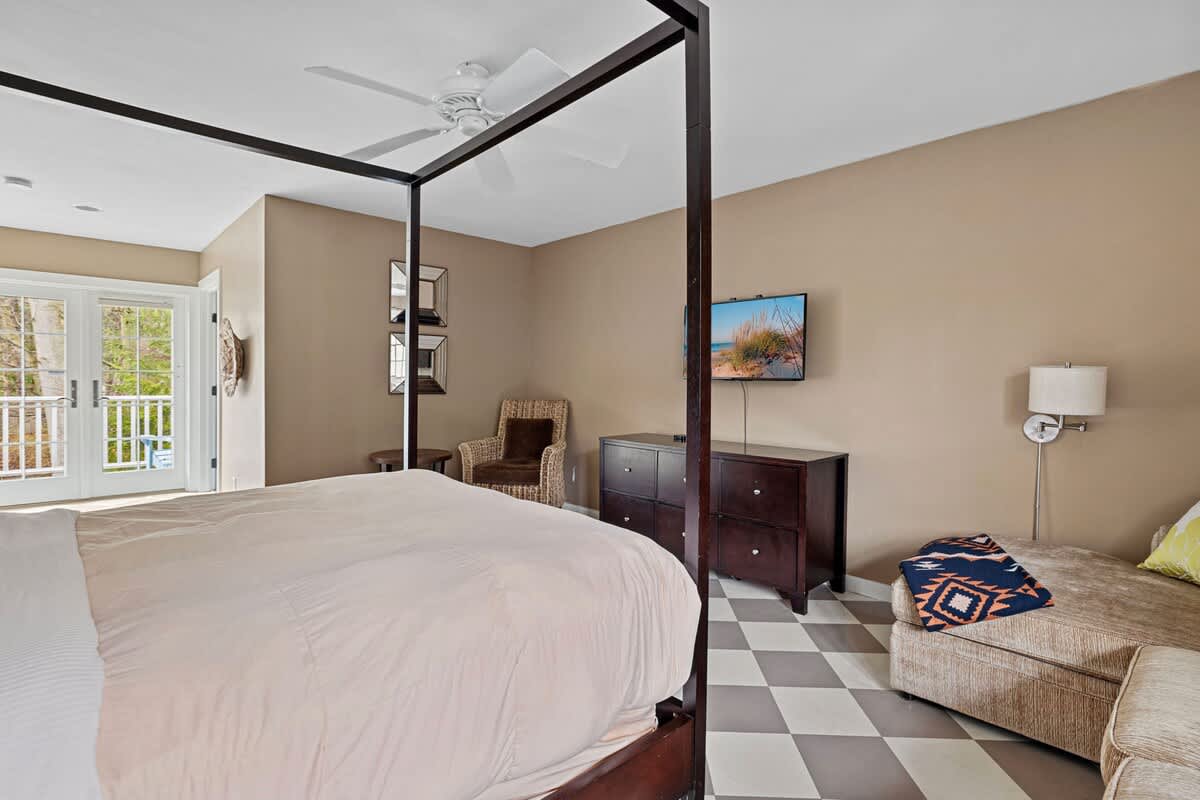 A spacious bedroom with light tan walls and a checkered floor, featuring a dark wood four-poster bed with light bedding, a wall-mounted TV, and a dresser. French doors lead to an outdoor area, and a ceiling fan is visible.