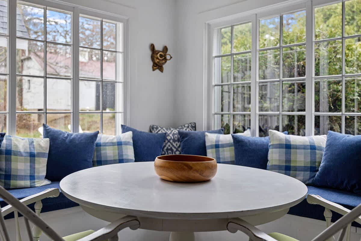 Bright breakfast nook with built-in blue cushioned bench seating and plaid pillows, surrounding a round white table, with large windows offering natural light.