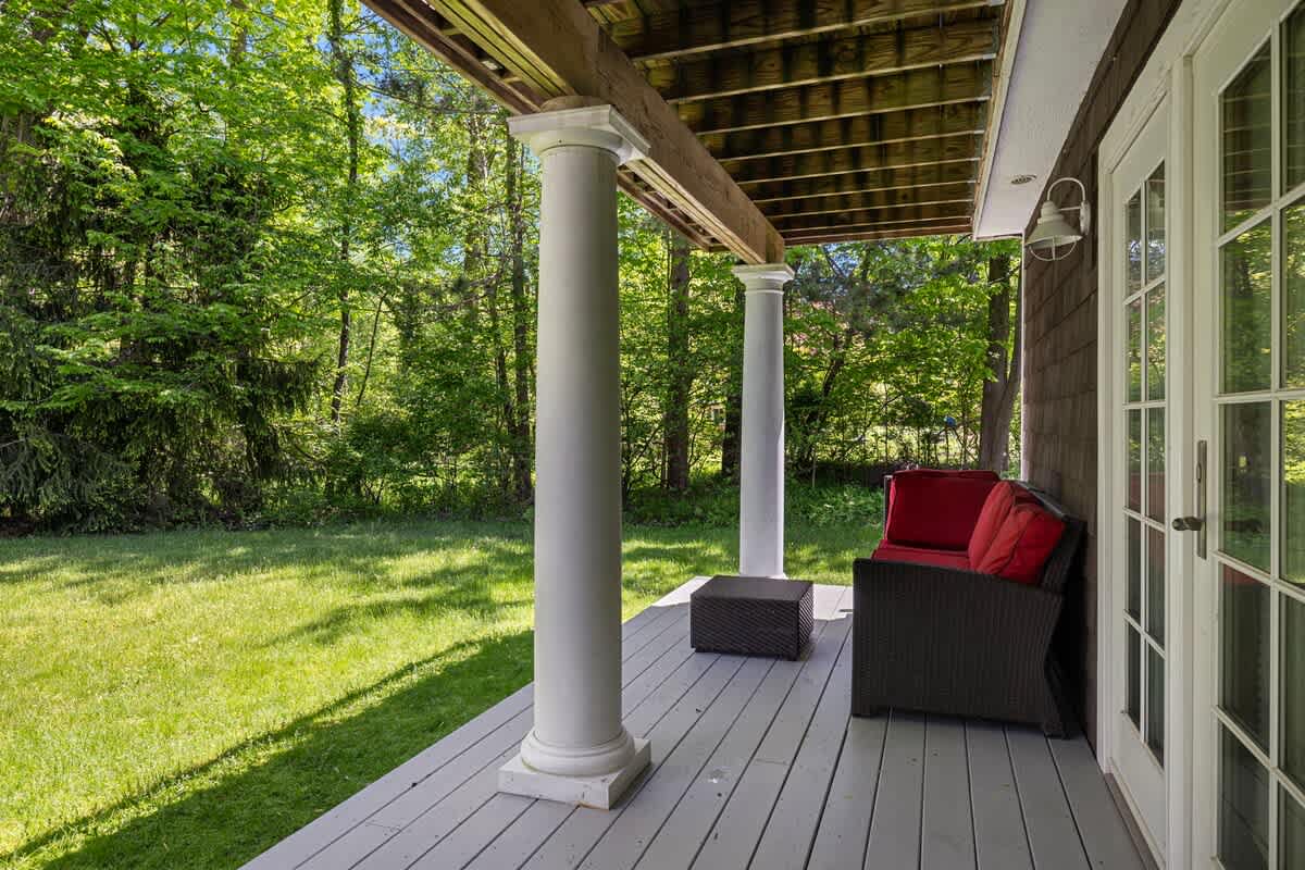 A covered outdoor deck with gray flooring and white columns, offering a view of a lush green yard and surrounding trees. A dark wicker sofa with red cushions provides seating.