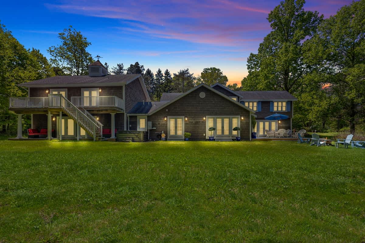 A large, multi-level house with dark shingle siding and white trim, featuring multiple decks and French doors, overlooking a vast green lawn under a vibrant sunset sky. Adirondack chairs and a fire pit are visible on the lawn.