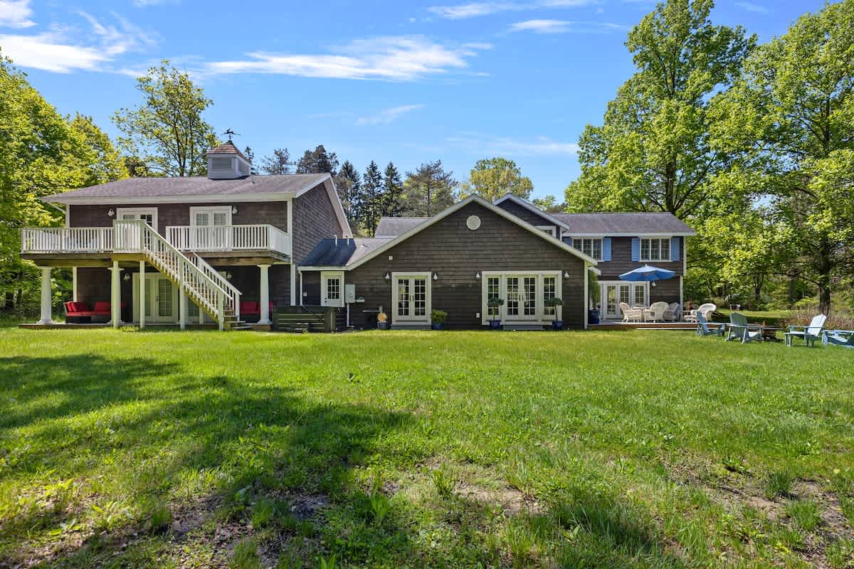 A sprawling green lawn extends behind a large, shingle-sided house with multiple wings and a deck. Outdoor seating areas, including a fire pit with Adirondack chairs, are visible, surrounded by mature trees under a blue sky.