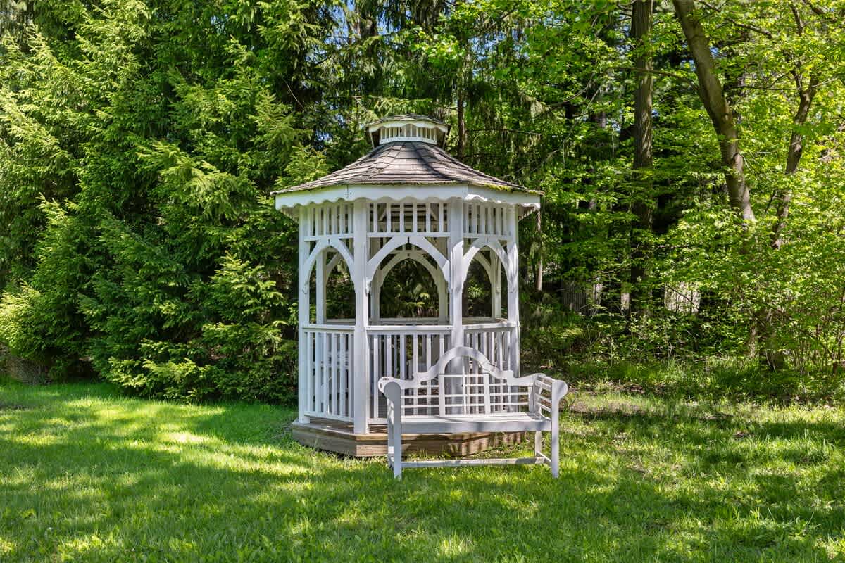 A picturesque white wooden gazebo with decorative arches and a shingled roof stands on a small wooden platform. A matching white garden bench is placed on the surrounding green lawn, with dense foliage in the background.