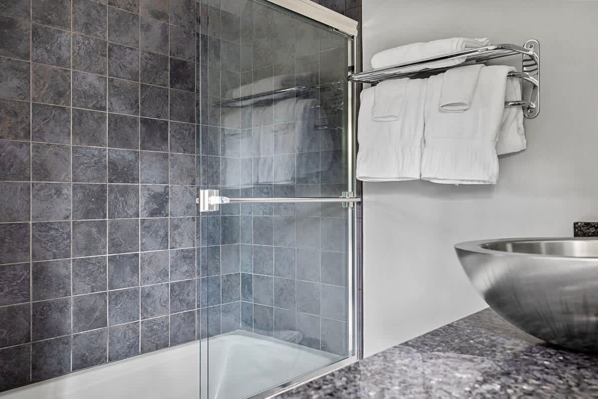 A modern bathroom featuring dark gray tiled walls in the shower/tub combo with a glass enclosure. A stainless steel vessel sink sits on a dark countertop, and white towels are neatly stacked on a wall-mounted rack.