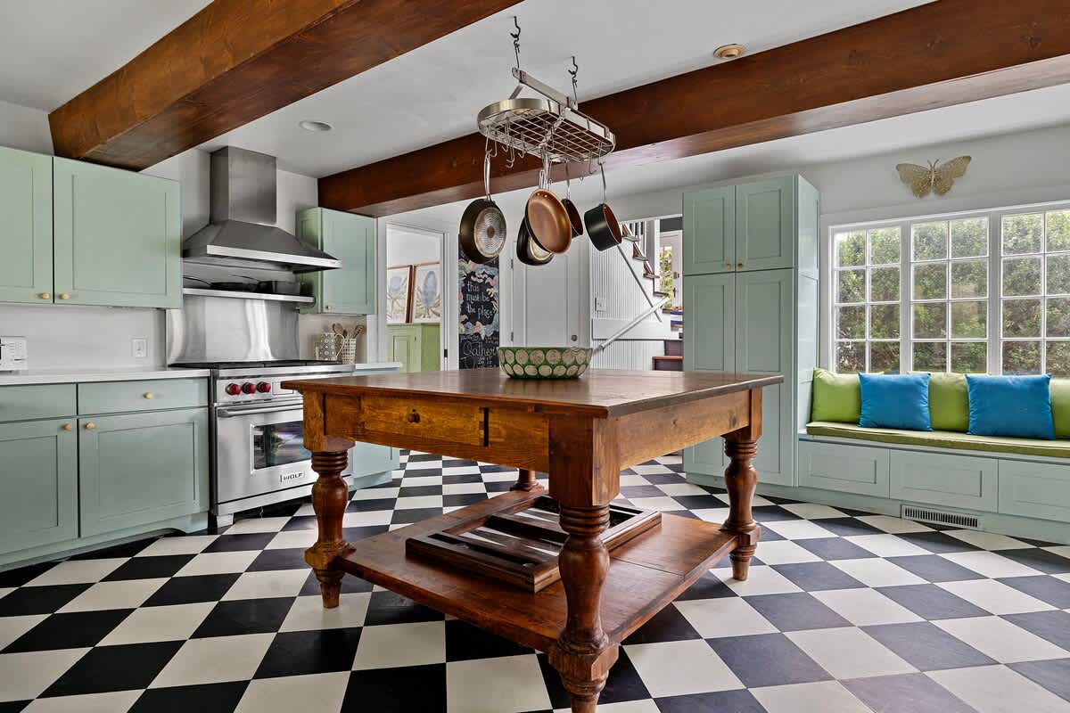 A spacious kitchen boasts light green cabinets, stainless steel appliances including a range and hood, and a large wooden island with a pot rack overhead. The floor features a striking black and white checkered pattern, and a built-in window seat with colorful cushions is visible.