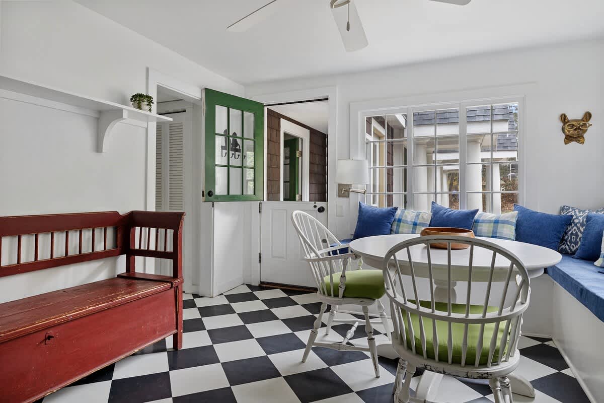 A charming breakfast nook with a round white table, white spindle-back chairs with green cushions, and a built-in bench with blue cushions. A green dutch door and black and white checkered floor add character to the space.